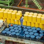 Workers managing blue and yellow barrels at a Houston transloading site with industrial piping and metal structures.