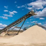 ndustrial site with a large pile of frac sand and conveyor belts under a blue sky with clouds.