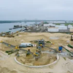 Industrial sand management facility with machinery and conveyor belts near a body of water, essential for oil and gas production well longevity.
