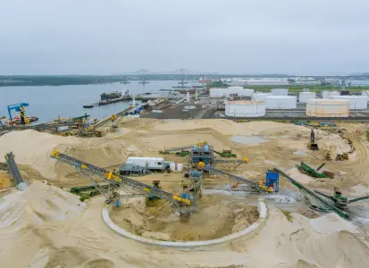 Industrial sand management facility with machinery and conveyor belts near a body of water, essential for oil and gas production well longevity.