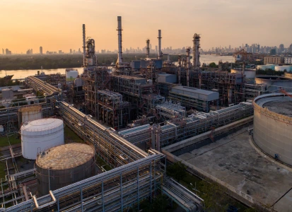 Aerial view of a U.S. oil refinery with pipelines and tanks near a river at sunset, illustrating midstream and upstream energy operations in 2025